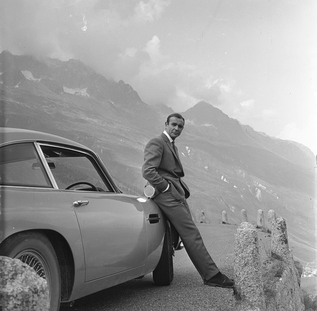 Sean Connery relaxes on the bumper of his Aston Martin DB5 during the filming of location scenes for Goldfinger in the Swiss Alps. Copyright Notice - © 1964 Danjaq, LLC and United Artists Corporation. All rights reserved. 1. SEAN CONNERY relaxes on the bumper of his Aston Martin DB5 © 1964 Danjaq, LLC and United Artists Corporation. - LR.jpg